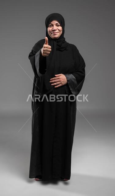 الوقوف باستقامة والنظر إلى الكاميرا، بورتريه لسيدة عربية خليجية سعودية مبتسمة محجبة كبيرة بالسن مبتسمة ترتدي العباءة السوداء تقف ترفع إصبع الإبهام نحو الأعلى بعلامة الجودة، إيماءات الإعجاب والرضا، صورة الجسم بالطول الكامل، خلفية رمادية.