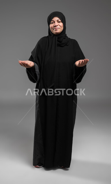 الشعور بالحيرة والجهل بأمر ما، التردد وانعدام المعرفة بخصوص شيء معين، بورتريه لسيدة كبيرة بالسن عربية سعودية خليجية محجبة ترتدي العباءة ترفع يديها بإيماءات عدم التأكد، صورة لكامل الجسم، خلفية رمادية.