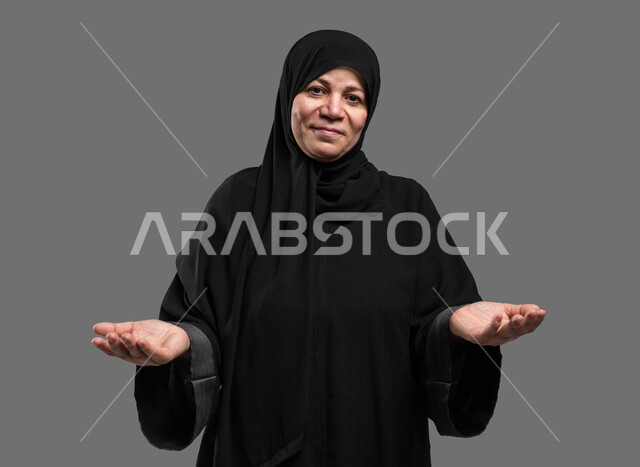 التردد وانعدام المعرفة بخصوص شيء معين، الشعور بالحيرة والجهل بأمر ما، بورتريه مقرب لسيدة كبيرة بالسن عربية سعودية خليجية محجبة ترتدي العباءة تنظر إلى الكاميرا وترفع يديها بإيماءات عدم التأكد، خلفية رمادية.