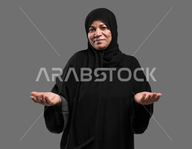 التردد وانعدام المعرفة بخصوص شيء معين، الشعور بالحيرة والجهل بأمر ما، بورتريه مقرب لسيدة كبيرة بالسن عربية سعودية خليجية محجبة ترتدي العباءة تنظر إلى الكاميرا وترفع يديها بإيماءات عدم التأكد، خلفية رمادية.