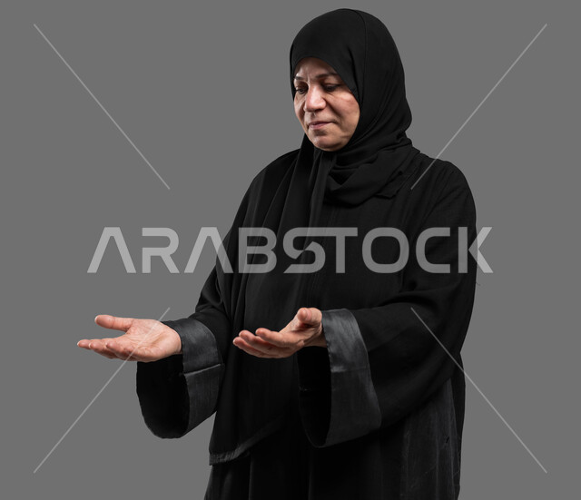 تقديم وعرض المساعدة للآخرين، بورتريه مقرب لسيدة كبيرة بالسن عربية سعودية خليجية محجبة مبتسمة ترتدي العباءة تفتح يديها لعرض شيء ما، رفع وحمل الأغراض والحاجات، خلفية رمادية.