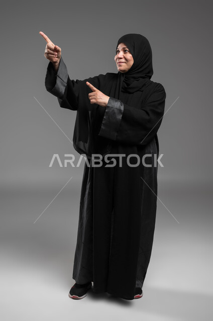 التفاعل وتحريك العناصر الهولوجرامية، الاستمتاع في محاكاة العالم الخيالي، النقر على الشاشة باستخدام حاسة اللمس، بورتريه لسيدة عربية سعودية خليجية كبيرة بالسن ترتدي العباءة والحجاب تنظر إلى الأعلى تشير بإصبعيها إلى شيء ما، صورة لكامل الجسم، خلفية رمادية.