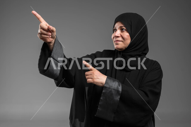 الاستمتاع في محاكاة العالم الخيالي، التفاعل وتحريك العناصر الهولوجرامية، النقر على الشاشة باستخدام حاسة اللمس، بورتريه مقرب لسيدة عربية سعودية خليجية كبيرة بالسن ترتدي العباءة والحجاب تنظر إلى الأعلى تشير بإصبعيها إلى شيء ما، خلفية رمادية.