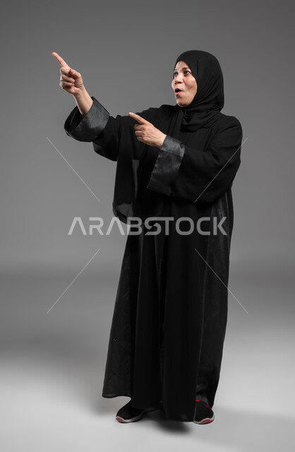 الاستمتاع في محاكاة العالم الخيالي، التفاعل وتحريك العناصر الهولوجرامية باستخدام حاسة اللمس، بورتريه لسيدة عربية سعودية خليجية محجبة كبيرة بالسن ترتدي العباءة تنظر إلى الأعلى تشير بإصبعيها إلى شيء ما بإيماءات الذهول، صورة الجسم بالكامل، خلفية رمادية.