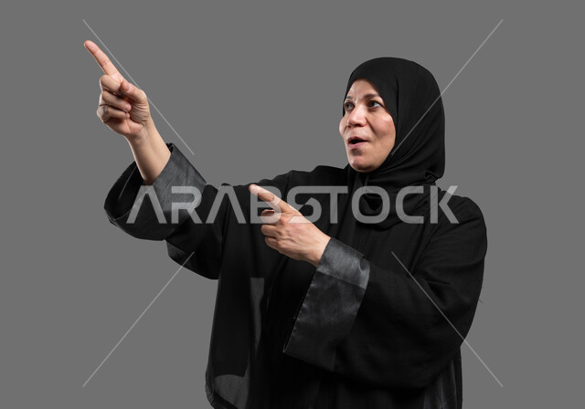 Tapping on the screen using the sense of touch, feeling the pleasure of simulating the imaginary world, interacting and moving holographic elements, a close-up portrait of an elderly Saudi Arabian Gulf woman wearing an abaya and veil, looking up, pointing her fingers at something with astonished gestures, gray background.