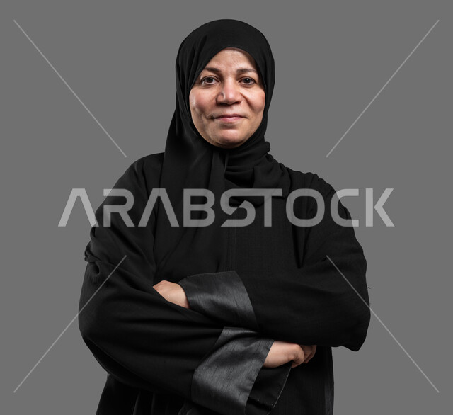 الوقوف بأيدي متقاطعة وثقة والنظر إلى الكاميرا، الاهتمام بالمظهر الخارجي، بورتريه مقرب لسيدة عربية خليجية سعودية محجبة كبيرة بالسن مبتسمة ترتدي العباءة السوداء تقف متكتفة اليدين بإيماءات السرور، خلفية رمادية.
