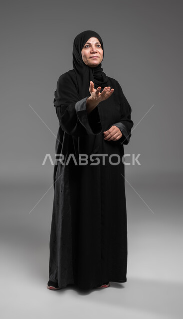 Feeling the pleasure of simulating the imaginary world, interacting with holographic elements, a portrait of an elderly veiled Saudi Arabian Gulf woman wearing an abaya, looking up and opening her hand to try to hold and capture something, full body image, gray background.
