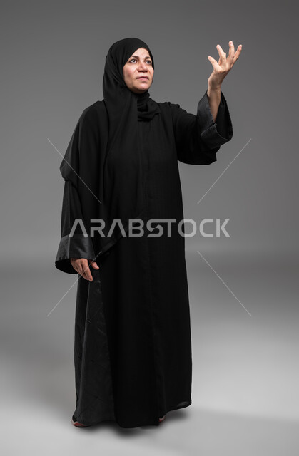 التساؤل والاستفسار لمعرفة موضوع مبهم، بورتريه لسيدة عربية سعودية خليجية كبيرة بالسن ترتدي العباءة والحجاب تنظر إلى الأعلى تشير بيدها للسؤال والاستفسار عن شيء ما بإيماءات الاستغراب، صورة لكامل الجسم، خلفية رمادية.