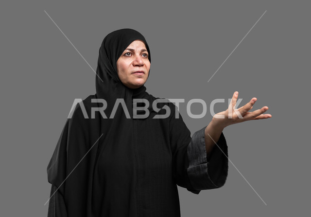 الاستفسار والتساؤل لمعرفة موضوع مبهم، بورتريه مقرب لسيدة عربية سعودية خليجية كبيرة بالسن ترتدي العباءة والحجاب تنظر إلى الأعلى تشير بيدها للسؤال والاستفسار عن شيء ما بإيماءات الاستغراب، خلفية رمادية.
