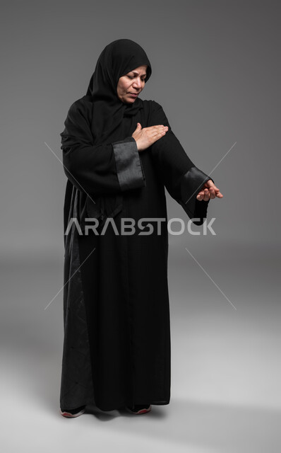 مشاكل صحية بسبب التقدم بالعمر، الحاجة إلى العلاج والمساعدة، المعاناة من الآلام والأوجاع العصبية والعضلية، بورتريه مقرب لسيدة عربية خليجية سعودية محجبة كبيرة بالسن ترتدي العباءة تضع يدها على ساعدها بإيماءات الشعور بالألم ، صورة الجسم بالكامل، خلفية رمادية.