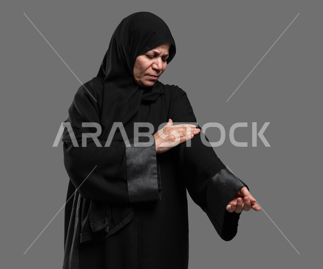 الحاجة إلى العلاج والمساعدة، مشاكل صحية وإصابات جسدية بسبب التقدم بالعمر، المعاناة من الآلام والأوجاع العصبية والعضلية، بورتريه مقرب لسيدة عربية خليجية سعودية محجبة كبيرة بالسن ترتدي العباءة تضع يدها على ساعدها بإيماءات الشعور بالألم والوجع، خلفية رمادية.