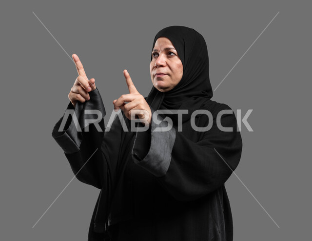 Enjoying the simulation of the imaginary world, interacting and moving holographic elements, tapping on the screen using the sense of touch, a close-up portrait of an elderly Saudi Arabian Gulf woman wearing an abaya and hijab, looking up and pointing with her fingers at something, gray background.