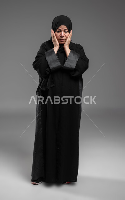 الشعور بالخيبة والانزعاج، الندم على شيء ما، بورتربه لسيدة عربية سعودية خليجية كبيرة بالسن ترتدي العباءة والحجاب تضع يديها على وجهها بإيماءات تدل على الحزن والإحباط، صورة لكامل الجسم، خلفية رمادية.