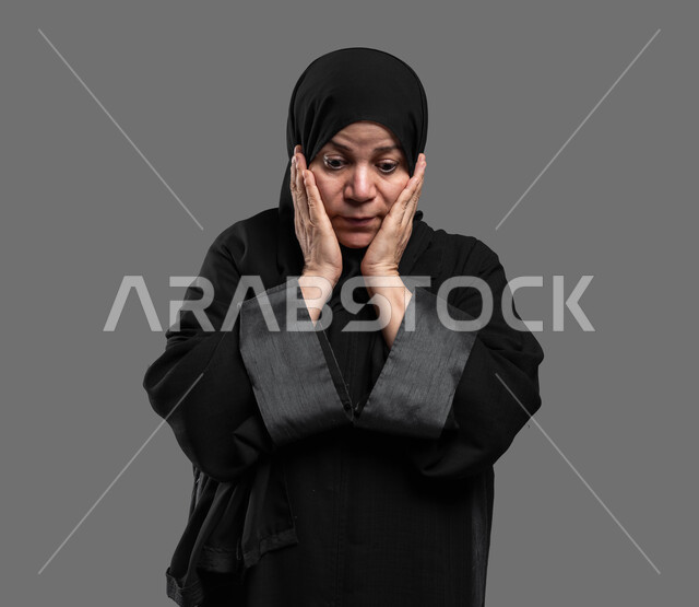 النظر إلى الأسفل بإيماءات الدهشة والاستغراب، التعجب من حصول أمر ما، بورتربه مقرب لسيدة عربية سعودية خليجية كبيرة بالسن ترتدي العباءة والحجاب تضع يديها على وجهها بإيماءات تدل على الصدمة والذهول، خلفية رمادية.