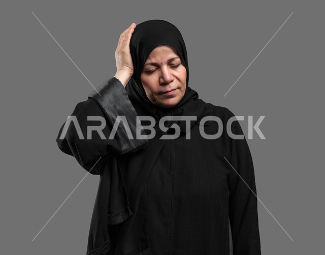 الحاجة للهدوء والراحة عند الكبار بالعمر، بورتريه مقرب لسيدة عربية سعودية خليجية كبيرة بالسن ترتدي العباءة والحجاب تضع يدها على رأسها بإيماءات تدل على الألم والتعب, الإحساس بوجع الرأس والصداع, آلام الشقيقة والصداع النصفي، خلفية رمادية.