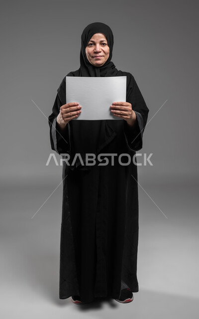 Displaying an empty square white board, using explanatory boards, advertisements and marketing presentations, a portrait of a veiled, smiling, elderly Saudi Arabian Gulf woman wearing an abaya, holding a white square mock-up in her hands, looking at the camera, a full-body portrait, gray background.
