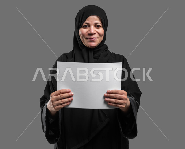 Displaying an empty square white board, using explanatory boards, advertisements and marketing presentations, a close-up portrait of a veiled, smiling, elderly Saudi Arabian Gulf woman wearing an abaya, holding a white square mock-up in her hand, looking at the camera with gestures of pleasure and joy, gray background.