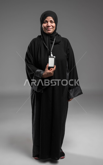 الوقوف بثقة والنظر إلى الكاميرا، ارتداء بادج شخصي، عرض بطاقة تعريفية، كرت أبيض فارغ مستطيل الشكل، بورتريه لسيدة عربية خليجية سعودية محجبة كبيرة بالسن مبتسمة ترتدي العباءة السوداء تضع هوية شخصية، صورة لكامل الجسم، خلفية رمادية.