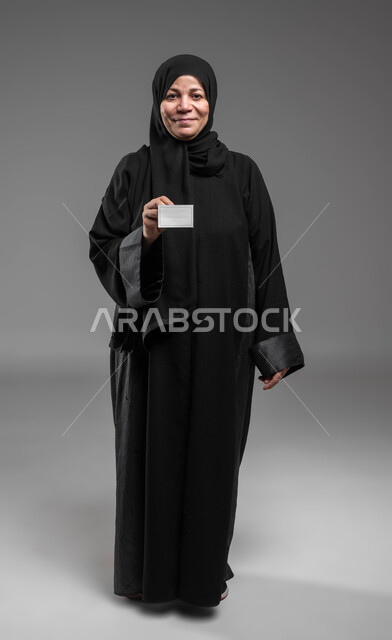 كرت أبيض فارغ مستطيل الشكل، كروت إعلانات وعروض تسويقية، عرض بطاقة تعريفية شخصية، بورتريه لامرأة عربية خليجية سعودية محجبة كبيرة بالسن مبتسمة ترتدي العباءة السوداء تحمل بيديها ورقة بيضاء فارغة، صورة الجسم بالكامل، خلفية رمادية.