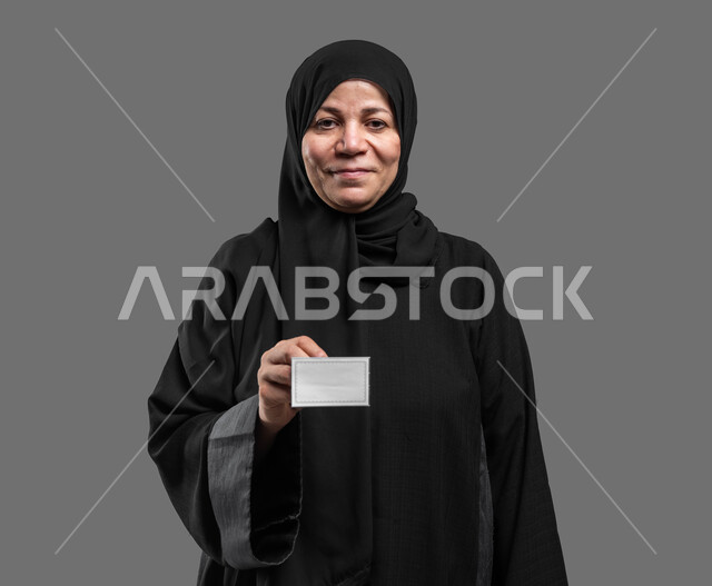 Displaying a personal identification card, a blank white rectangular card, advertising cards and marketing offers, a close-up portrait of a veiled, elderly Saudi Arabian Gulf woman, smiling, wearing a black abaya, looking at the camera, holding a blank white sheet of paper in her hands, a gray background.