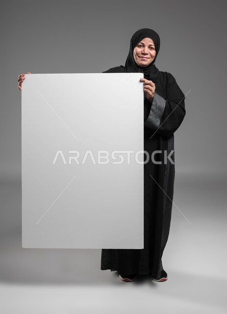 عرض إعلانات وعروض تسويقية، استخدام لوحات توضيحية ووسائل تعليمية، موك اب مربع أبيض كبير، بورتريه لامرأة عربية خليجية سعودية محجبة كبيرة بالسن مبتسمة ترتدي العباءة السوداء تحمل بيديها لوحه بيضاء مربعه فارغة تنظر إلى الكاميرا، صورة الجسم بالكامل، خلفية رمادية.