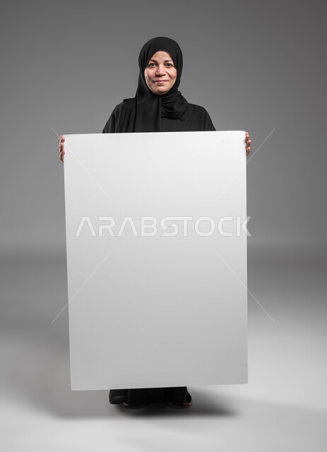 موك اب مربع أبيض كبير، عرض إعلانات وعروض تسويقية، استخدام لوحات توضيحية ووسائل تعليمية، بورتريه لامرأة عربية خليجية سعودية محجبة كبيرة بالسن مبتسمة ترتدي العباءة السوداء تحمل بيديها لوحه بيضاء مربعه فارغة تنظر إلى الكاميرا، صورة الجسم بالكامل، خلفية رمادية.