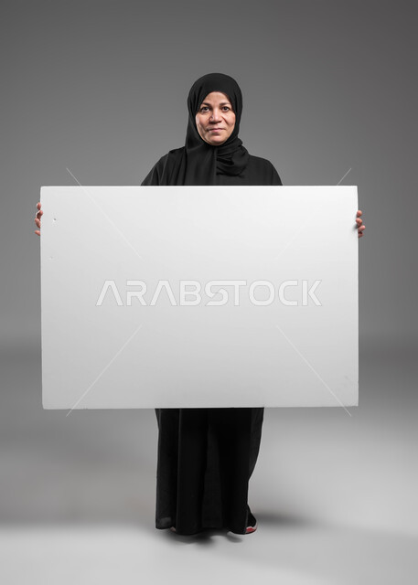 عرض إعلانات وعروض تسويقية، استخدام لوحات توضيحية ووسائل تعليمية، موك اب مربع أبيض كبير، بورتريه لامرأة عربية خليجية سعودية محجبة كبيرة بالسن مبتسمة ترتدي العباءة السوداء تحمل بيديها لوحه بيضاء مربعه فارغة تنظر إلى الكاميرا، صورة الجسم بالكامل، خلفية رمادية.