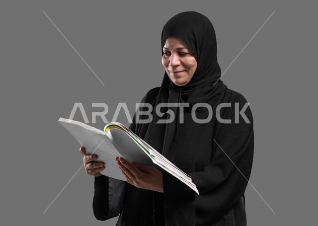 قضاء وقت الفراغ بالتثقيف والتعلم، مفهوم محو الأمية، هواية المطالعة وتثقيف النفس، بورتريه مقرب لسيدة كبيرة بالسن عربية سعودية خليجية محجبة مبتسمة ترتدي العباءة تقرأ كتاب بإيماءات التمعن والتركيز، خلفية رمادية.