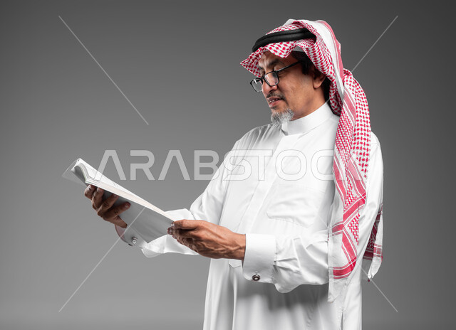 هواية المطالعة وتثقيف النفس، قضاء وقت الفراغ بالتثقيف والتعلم، مفهوم محو الأمية، بورتريه مقرب لرجل كبير بالسن عربي سعودي خليجي يرتدي الشماغ والثوب التقليدي والنظارة الطبية يقرأ كتاب بإيماءات التمعن والتركيز، خلفية رمادية.
