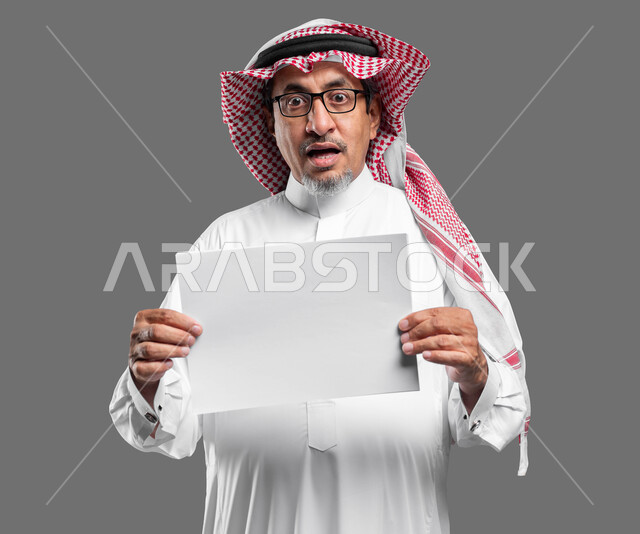 لوحه بيضاء مربعه فارغة، استخدام لوحات توضيحية تعريفية، إعلانات وعروض تسويقية، بورتريه لرجل عربي خليجي سعودي كبير بالسن يرتدي الشماغ  والثوب التقليدي ونظارة طبية يحمل بيده موك اب مربع أبيض ينظر إلى الكاميرا بايماءات الدهشة، صورة الجسم بالكامل، خلفية رماديه