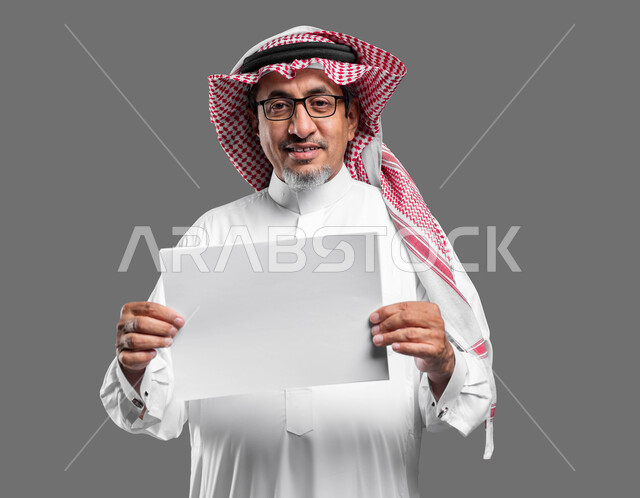 لوحه بيضاء مربعه فارغة، استخدام لوحات توضيحية تعريفية، إعلانات وعروض تسويقية، بورتريه مقرب لرجل عربي خليجي سعودي كبير بالسن مبتسم يرتدي الشماغ  والثوب التقليدي ونظارة طبية يحمل بيده موك اب مربع أبيض ينظر إلى الكاميرا، صورة الجسم بالكامل، خلفية رماديه