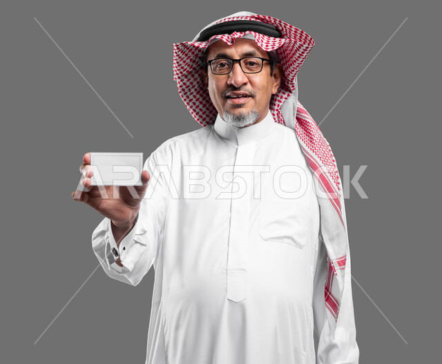 كرت أبيض مستطيل فارغ، استخدام بطاقات توضيحية تعريفية، كروت إعلانات وعروض تسويقية، بورتريه مقرب لرجل عربي خليجي سعودي كبير بالسن مبتسم يرتدي الشماغ  والثوب التقليدي ونظارة طبية يحمل بيده موك اب مربع أبيض ينظر إلى الكاميرا، خلفية رماديه