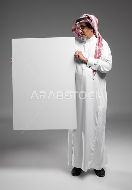 A large white square mockup, a portrait of an elderly Saudi Gulf Arab man, smiling, wearing a shemagh, a traditional thobe, and prescription glasses, holding a blank square white board in his hand, looking at the camera, advertisements and marketing presentations, using explanatory panels and educational aids, full body image, gray background.