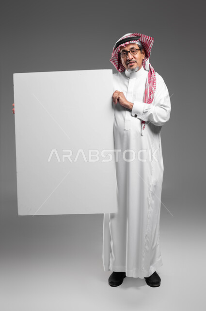 موك اب مربع أبيض كبير، بورتريه لرجل عربي خليجي سعودي كبير بالسن مبتسم يرتدي الشماغ  والثوب التقليدي ونظارة طبية يحمل بيده لوحه بيضاء مربعه فارغة ينظر إلى الكاميرا، إعلانات وعروض تسويقية، استخدام لوحات توضيحية ووسائل تعليمية، صورة الجسم بالكامل، خلفية رماديه