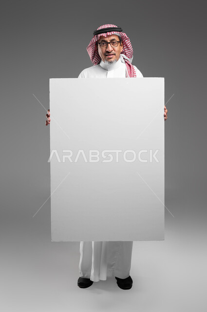 Advertisements and marketing offers, use of explanatory panels and educational materials, a large white square mockup, a portrait of an elderly Saudi Arabian Gulf man, smiling, wearing a shemagh, a traditional thobe, and medical glasses, holding a blank square white board in his hand, looking at the camera, full body portrait, gray background.