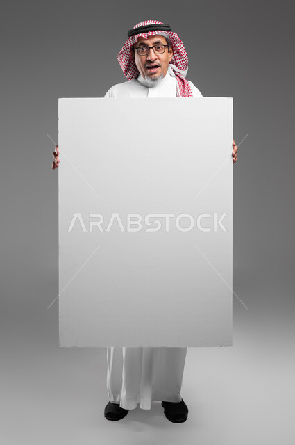 استخدام لوحات توضيحية تعريفية، إعلانات وعروض تسويقية، موك اب مربع أبيض كبير، بورتريه لرجل عربي خليجي سعودي كبير بالسن يرتدي الشماغ  والثوب التقليدي ونظارة طبية يحمل بيده لوحه بيضاء مربعه فارغة ينظر إلى الكاميرا بايماءات الدهشة، صورة الجسم بالكامل، خلفية رماديه