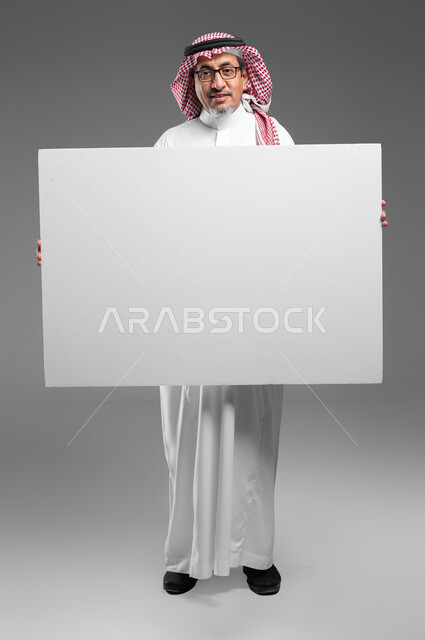 إعلانات وعروض تسويقية، استخدام لوحات توضيحية ووسائل تعليمية، موك اب مربع أبيض كبير، بورتريه لرجل عربي خليجي سعودي كبير بالسن مبتسم يرتدي الشماغ  والثوب التقليدي ونظارة طبية يحمل بيده لوحه بيضاء مربعه فارغة ينظر إلى الكاميرا، صورة الجسم بالكامل، خلفية رماديه