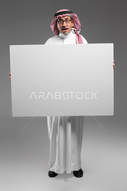 استخدام لوحات توضيحية تعريفية، إعلانات وعروض تسويقية، موك اب مربع أبيض كبير، بورتريه لرجل عربي خليجي سعودي كبير بالسن يرتدي الشماغ  والثوب التقليدي ونظارة طبية يحمل بيده لوحه بيضاء مربعه فارغة ينظر إلى الكاميرا بايماءات الدهشة، صورة الجسم بالكامل، خلفية رماديه