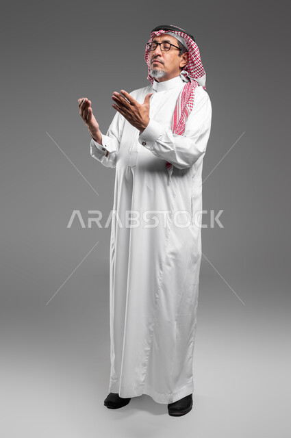 مفهوم الدعاء والترجي والطلب، الإجتهاد في العباده والتقرب الى الله في شهر رمضان المبارك, بورتريه لرجل عربي سعودي خليجي كبير بالسن يرتدي الشماغ والثوب التقليدي ونظارة طبية يرفع يديه ويدعو الله بإيماءات تدل على التوسل والتضرع، صورة الجسم بالكامل، خلفية رمادية
