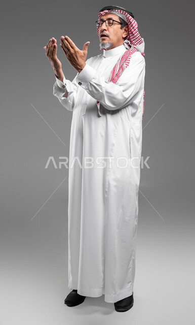 الإجتهاد في العباده والتقرب الى الله في شهر رمضان المبارك, مفهوم الدعاء والترجي والطلب، بورتريه لرجل عربي سعودي خليجي كبير بالسن يرتدي الشماغ والثوب التقليدي ونظارة طبية يرفع يديه ويدعو الله بإيماءات تدل على التوسل والتضرع، صورة الجسم بالكامل، خلفية رمادية