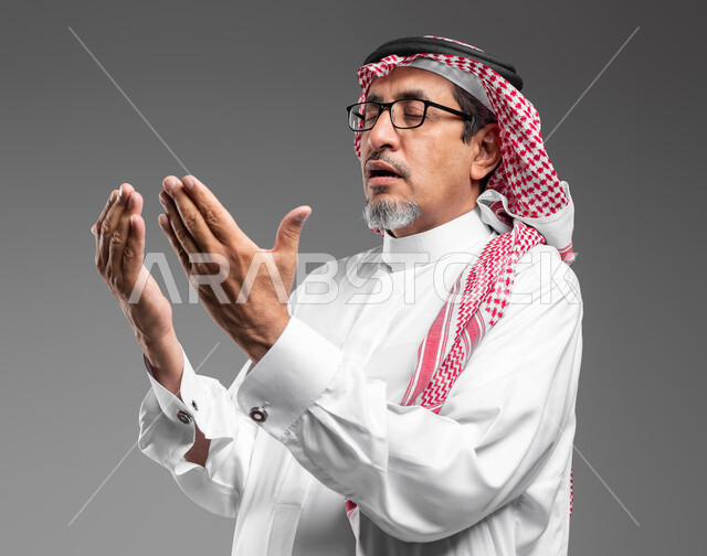 Diligence in worship and getting closer to God in the blessed month of Ramadan, the concept of supplication, hope and request, a close-up portrait of an elderly Saudi Arabian Gulf man wearing a shemagh, traditional dress, and medical glasses, raising his hands and calling upon God with gestures indicating pleading and supplication, gray background
