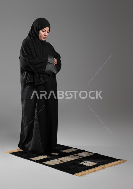 أهمية الالتزام بالصلاة على وقتها, التقرب من الله تعالى بالتزام أوامره, بورتريه لإمرأة عربية خليجية سعودية كبيرة بالسن ترتدي الحجاب والعباءة السوداء تقوم بأداء فروض الصلوات الخمس, مفهوم أركان الإسلام, إسلاميات وعبادة, صورة الجسم بالطول الكامل، خلفية رمادية