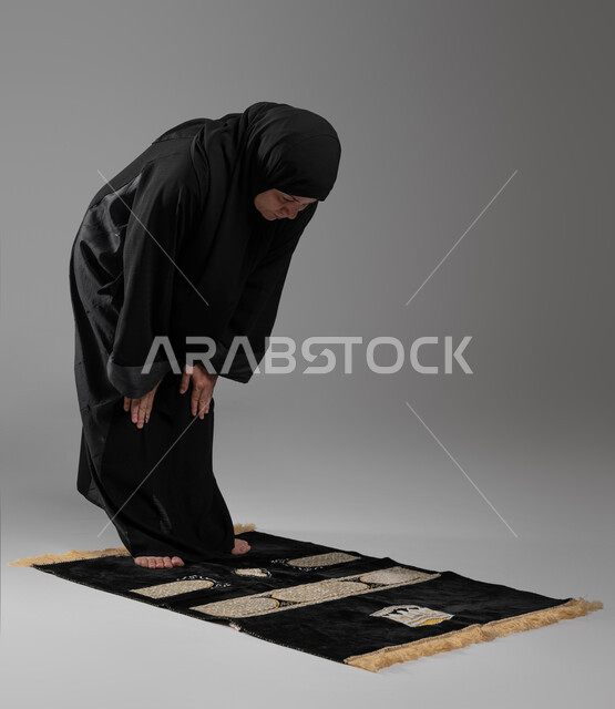 الركوع والسجود والخشوع لله، التقرب من الله تعالى بالتزام أوامره, بورتريه لإمرأة عربية خليجية سعودية كبيرة بالسن ترتدي الحجاب والعباءة السوداء تقوم بأداء فروض الصلوات الخمس, أهمية الالتزام بالصلاة على وقتها, إسلاميات وعبادة, صورة الجسم بالطول الكامل، خلفية رمادية