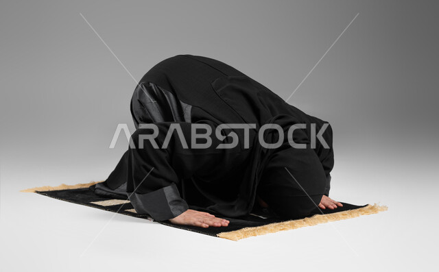 أهمية الالتزام بالصلاة على وقتها, التقرب من الله تعالى بأداء الفرائض والتزام أوامره, بورتريه لإمرأة عربية خليجية سعودية كبيرة بالسن ترتدي الحجاب والعباءة السوداء تسجد لله تعالى, مفهوم أركان الإسلام, إسلاميات وعبادة, خلفية رمادية