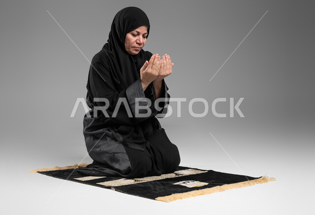 Performing the obligatory prayers on time, getting closer to God through diligence in worship and maintaining prayer, a portrait of an elderly Saudi Arabian Gulf woman wearing the hijab and black abaya, sitting on the prayer rug, raising her hands and calling upon God, gestures indicating pleading and pleading, gray background.
