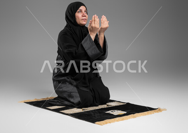إيماءات تدل على التوسل والتضرع، أداء فروض الصلاة على وقتها, التقرب الى الله من خلال الإجتهاد في العبادة والحفاظ على الصلاة، بورتريه لإمرأة عربية سعودية خليجية كبيرة بالسن ترتدي الحجاب والعباءة السوداء تجلس على سجادة الصلاة ترفع يديها وتدعو الله، خلفية رمادية