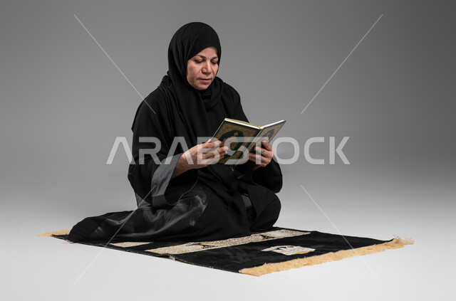 مشاعر الطمأنينه والخشوع, تلاوة وقراءة القرآن الكريم، بورتريه لإمرأة عربية سعودية خليجية ترتدي الحجاب والعباءة السوداء تجلس على سجادة الصلاة تحمل بيدها المصحف الشريف, الإكثار من ذكر الله تعالى والإجتهاد في العباده, التقرب الى الله في شهر رمضان, خلفية رمادية