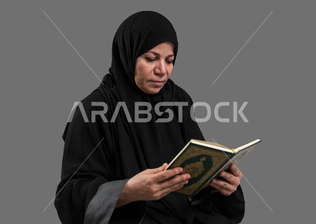 الشعور بالخشوع والطمأنينه, تلاوة وقراءة القرآن الكريم، بورتريه مقرب لإمرأة عربية سعودية خليجية ترتدي الحجاب والعباءة السوداء تجلس على سجادة الصلاة تحمل بيدها المصحف الشريف, الإكثار من ذكر الله تعالى والإجتهاد في العباده, التقرب الى الله في شهر رمضان, خلفية رمادية