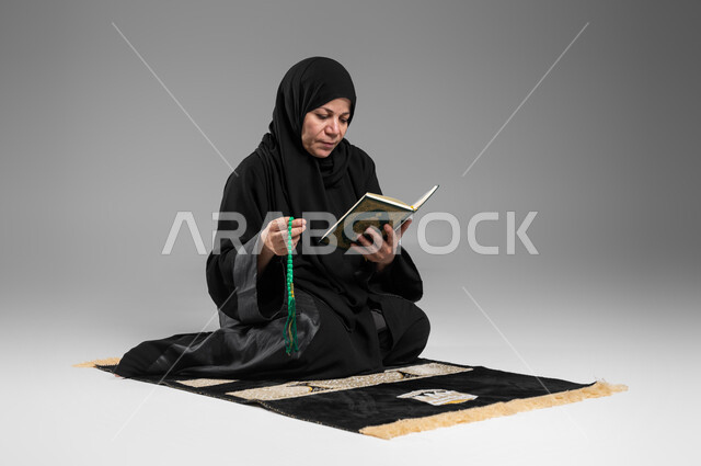 اسلاميات وعبادة، الإكثار من ذكر الله تعالى والإجتهاد في العباده, قراءة وتلاوة القرآن الكريم، بورتريه لإمرأة عربية سعودية خليجية كبيرة بالسن ترتدي الحجاب والعباءة السوداء تحمل بيدها المصحف الشريف والسبحة, التقرب الى الله في شهر رمضان المبارك, خلفية رمادية