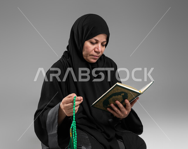 الإكثار من ذكر الله تعالى والإجتهاد في العباده, قراءة وتلاوة القرآن الكريم، بورتريه مقرب لإمرأة عربية سعودية خليجية كبيرة بالسن ترتدي الحجاب والعباءة السوداء تحمل بيدها المصحف الشريف والسبحة, التقرب الى الله في شهر رمضان المبارك, اسلاميات وعبادة، خلفية رمادية